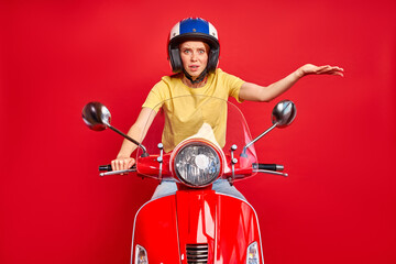 furious caucasian woman misunderstands situation on road while driving a motorbike, stands at a loss, wearing helmet isolated on red background