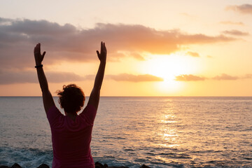Fototapeta premium Young woman wearing a pink top facing the ocean and exercising with arms raised, relaxing or doing yoga, enjoying quiet time and viewing a spectacular sunset, Palm Mar, Tenerife, Canary Islands, Spain