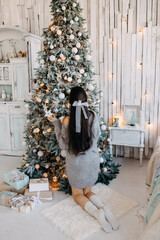 Fototapeta na wymiar Young brunette woman with a bow knot, wearing a cozy sweater, decorating Christmas tree at home.