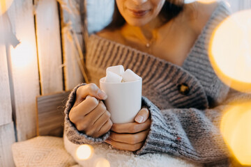 Fototapeta na wymiar Woman holding a cup of cocoa with marshmallows, wearing a cozy sweater at home.
