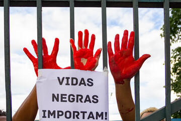 Três mãos pintadas com tinta vermelha por trás de grade de portão. Foto feita em manifestação...