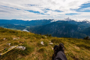 Sitting on top of the mountain Planina Blato in the Triglav National Park in Slovenia with panoramic view over the Lake Bohinj and the village Stara Fužina with outstretched legs in foreground
