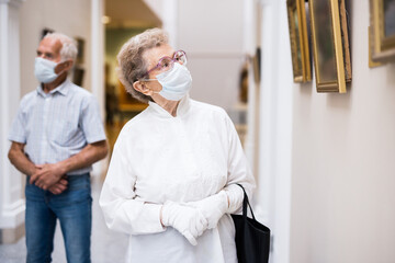 elderly European woman  in mask protecting against covid examines paintings on display in hall of art museum