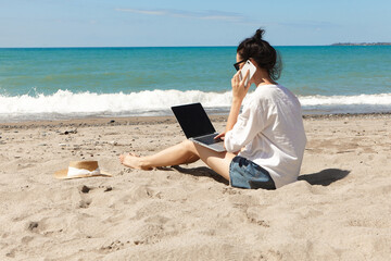 Young woman using laptop computer and talking on the phone on a beach. Vacation lifestyle...
