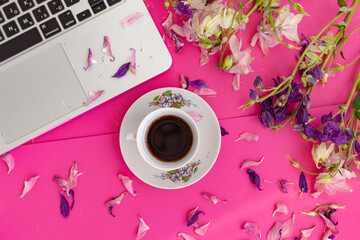 Home office desk. Female workspace with laptop and coffee mug, flowers and petals on pink...