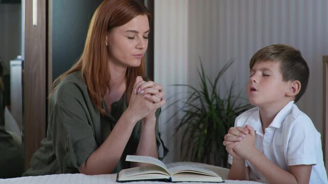 Believing Family, Female Parent With Her Male Child Kneel By Bed With Folded Hands And Pray To God Before Going To Bed In Room