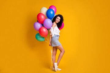 Fototapeta na wymiar Full size profile photo of cool cute brown curly girl hold many balloons behind back wear top skirt isolated on yellow color background