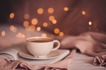 Tasty fresh cup of coffee on open paper book over Christmas lights close up. Good morning. Breakfast time. Winter holiday season.