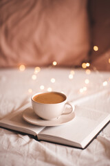 Mug of fresh tasty coffee on open paper book in bed over Christmas lights close up. Good morning. Winter holiday season. Breakfast time.