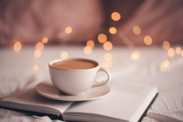 Cup of coffee stayin on open pepaer book over glowing Christmas lights close up. Good morning. Breakfast time. Xmas. Winter holiday time.