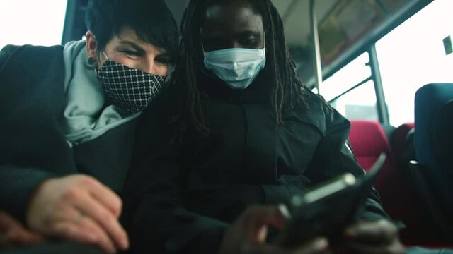 A black man in a mask and a white woman in a mask are looking at the phone screen on the bus. They are cheating. Exchange of information using the mobile Internet. Ease of showing than talking.