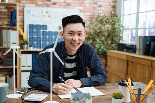 Attentive engineer. Smart responsible skilled architect working on wooden table with windmill and miniature house model creative eco friendly renewable building. handsome guy worker face camera smile