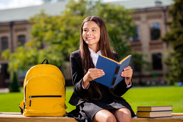 Photo of positive smart schoolgirl sit bench read book near bag wear black uniform white shirt...