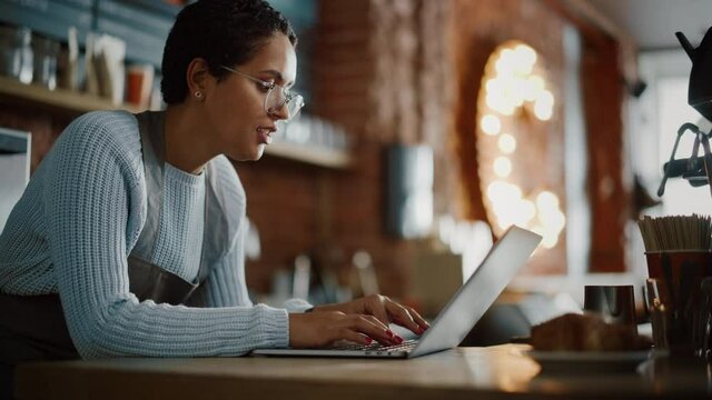 Young and Beautiful Latina Coffee Shop Owner is Working on Laptop Computer and Checking Inventory in a Cozy Cafe