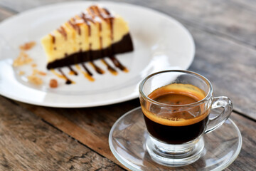 Close-up view, Espresso in shot glass with blurred cheesecake background on old wooden table.