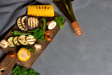 Grilled organic vegetables on a wooden cutting board and a bottle of wine on a gray background. Fried cornmeal, eggplant, mushrooms, tomatoes and spinach leaves. Grill menu