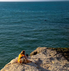 Fototapeta na wymiar Young woman with her dog sitting on a cliff. Sunny day blue ocean waves, tranquility, peace, joy. Spending time with your pet 
