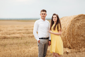 Photo of joyful couple man and woman walking through golden field with bunch of haystacks and hugging together during sunny day