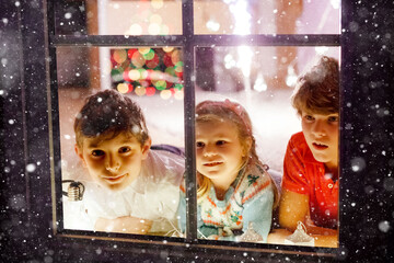 Three cute children sitting by window on Christmas eve. Two school kid boys and toddler girl, siblings looking outdoor and dreaming. Family happiness on traditional holiday