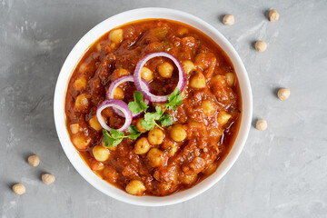 Top closeup view of chole masala or chana an indian street food made of chickpeas, tomatoes and cumin decorated with onion rings and parsley in bowl on gray concrete table. Image with copy space