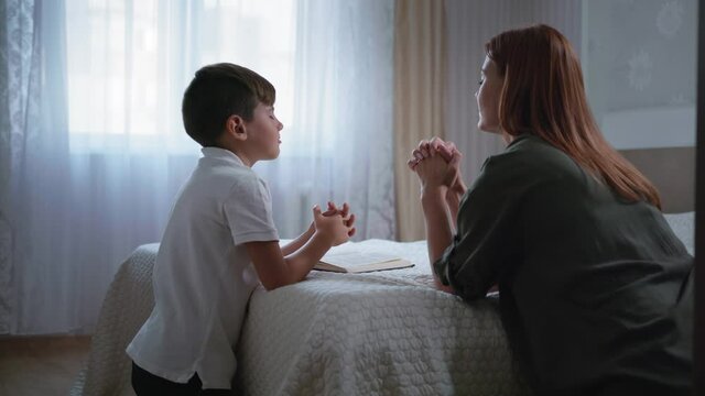 christianity, young woman together her son with closed eyes and folded hands with faith and hope in her heart praying to God kneeling by bed in room, religion concept
