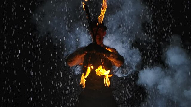 A male fire tamer in a spectacular close up fire show against a dark studio background among raindrops. A silhouette of a man in smoke waves flaming torches. Slow motion. Close up.
