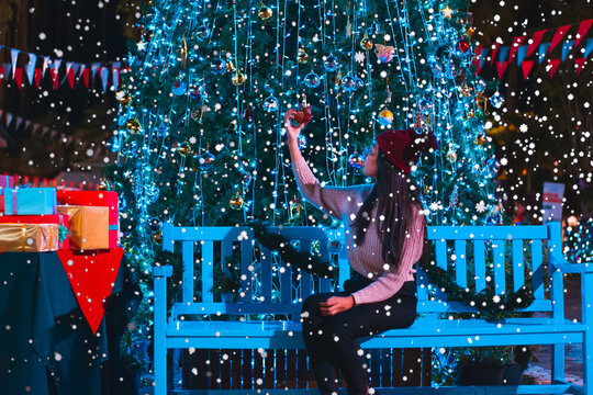 Young Woman Touching Decorations Hanging On Christmas Tree At Night During Snowfall