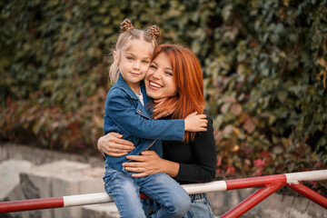 Fototapeta premium Stylish red-haired mother and her daughter are talking warmly. Happy child and her mom on the street. Successful single mother with her daughter for a walk. Warm family relationship