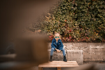 A cute little girl is sitting in sunglasses on a stone fence. Stylish child in a blue jacket. Casual style, fashion for children, fashionable suit, happy childhood