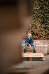 A cute little girl is sitting in sunglasses on a stone fence. Stylish child in a blue jacket. Casual style, fashion for children, fashionable suit, happy childhood