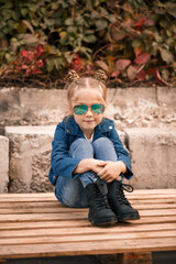 A cute little girl is sitting in sunglasses on a stone fence. Stylish child in a blue jacket. Casual style, fashion for children, fashionable suit, happy childhood