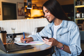 Beautiful brunette girl uses gadgets for telecommuting from cafe, holds smartphone in her hand. Online retraining
