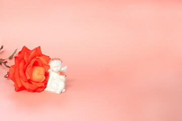 figure of a white angel on a pink background with a red flower in his hands, next lies an orange rose.