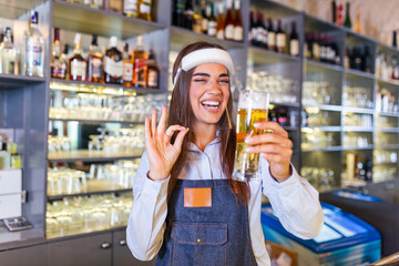 Bartender with face shield,covid-19 protection, serving a draft beer at the bar counter during coronavirus pandemic, showing OK sign,shelves full of bottles with alcohol on the background