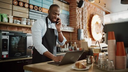 Black African American Coffee Shop Employee Accepts a Pre-Order on a Mobile Phone Call and Writes it Down on Laptop Computer in a Cafe. Restaurant Manager Browsing Internet and Talking on Smartphone. - Powered by Adobe