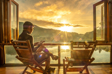 young man reading book near window and watching lake view at coffee shop in the morning sunrise,...