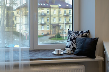 Obraz premium Cozy part of the kitchen in an old residential house with a view of the city through the window. The windowsill covered with a gray fabric overlay, on it two black-white pillows and cups of coffee