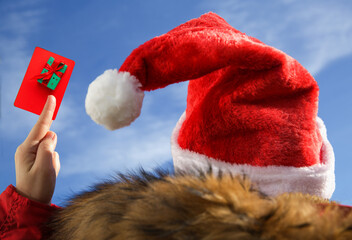 Close-up of a female hand holding a gift card with a picture of a gift against the blue sky.in Santa hat and red jacket. gift or discount concept