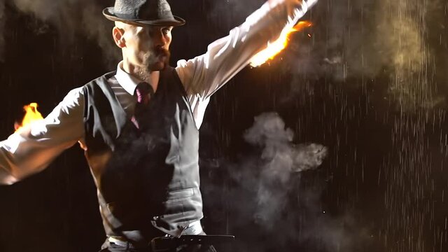 An exciting show of fire under streams of rain in a dark studio. A stylish man demonstrates a light show and rotates burning torches. Close up silhouette. Slow motion.