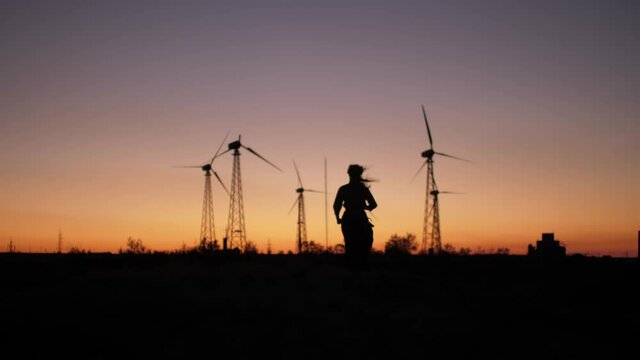 Slow-motion scene of pretty girls silhouette runs trough the hills near Wind Turbine in sunset light. Beautiful summer colors of childhood.