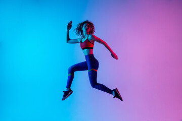 High jump. Beautiful woman in bright sportwear isolated on gradient pink-blue background in neon light. Athletic and graceful. Modern sport, action, motion, youth concept. Sportive female practicing