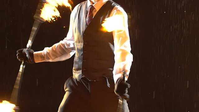 An exciting show of fire under streams of rain in a dark studio. A stylish man demonstrates a light show and rotates burning torches. Close up silhouette. Slow motion.