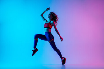 Run and jump. Young sportive woman training isolated on gradient blue-pink background in neon...
