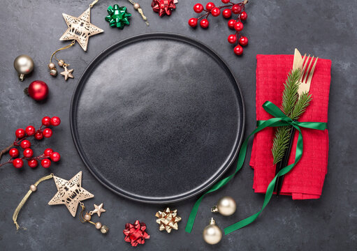 Christmas table setting with black ceramic plate, fir tree branch and gold and red accessories on black stone background. Top view. Copy space