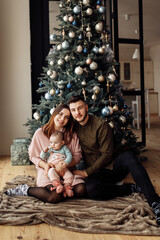 Fototapeta na wymiar Lovely parents with cute kid sit near Christmas tree at home, smile, caring mom hold little daughter in arms, family enjoy winter holidays together, New Year celebration concept