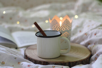 Enamel mug with hot drink and cinnamon stick, lit candle and open book on the bed. Selective focus, bokeh lights.