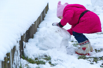 A girl rolls a big lump for a snowman. She's wearing a red coat. boots, mittens
