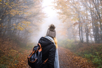 Woman with backpack and knit hat hiking in foggy forest. Tourist looking for adventure in nature