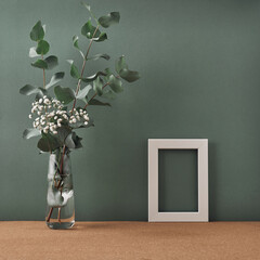 Frame and vase with white flowers and eucalyptus on a dark green background. Mock up, copy space. Folk.