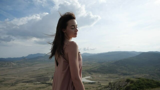 A video portrait of a slender girl looking at the camera in a pink dress enjoying the sun's rays falling on her face, the wind flutters her hair against the backdrop of the mountainous terrain